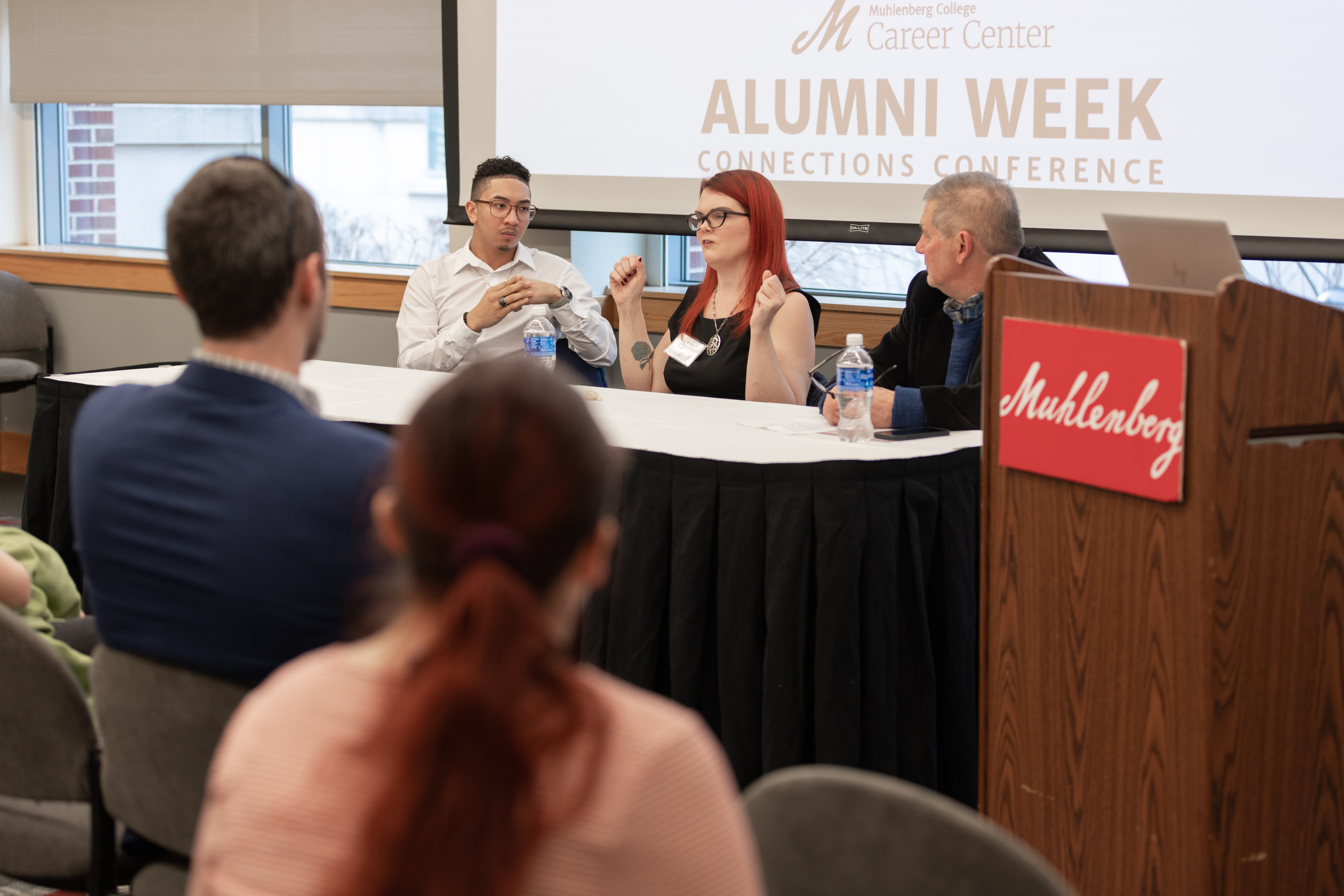 Three adults sit in front of a screen that says Alumni Week having a discussion in front of an audience